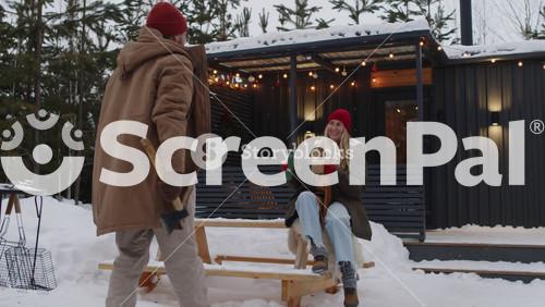 Full Shot Of Man Chopping Logs With Axe In Snowy Front Yard And Coming Up To Smiling Wife Who Giving Cup Of Hot Tea, Spending Christmas In Countryside Together